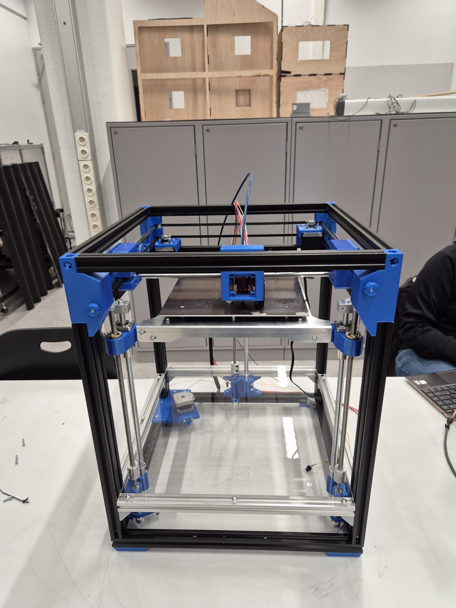 The finished CoreXY 3D printer, a black aluminium-extrusion cube with blue printed brackets, on a lab table
