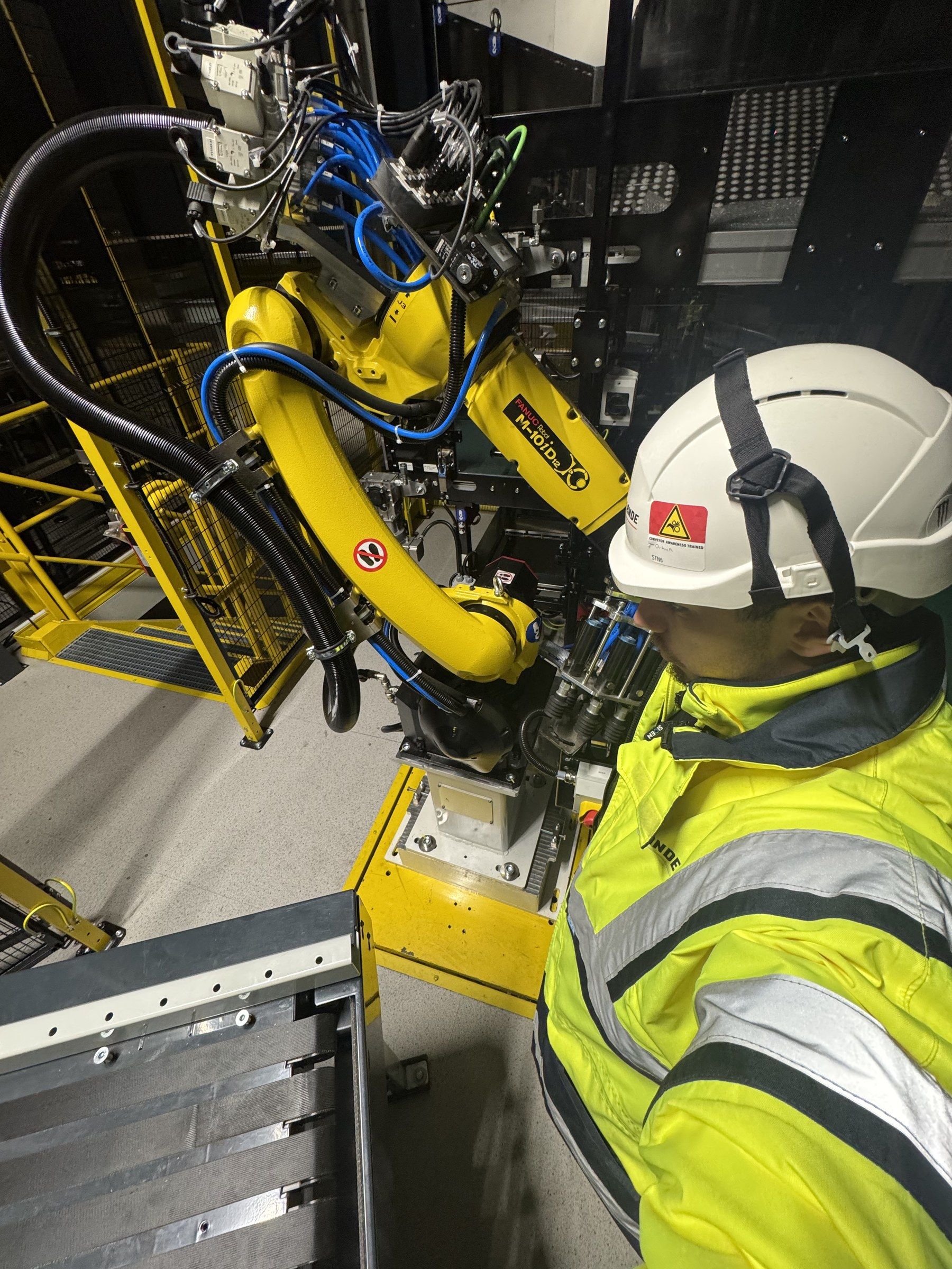 Orhun in a hard hat and hi-vis next to a yellow industrial robot arm